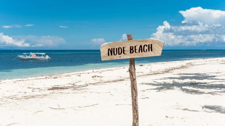 Blick auf einen weißen Sandstrand, türkisfarbenes Meer und ein Schild mit der Aufschrift Nude Beach